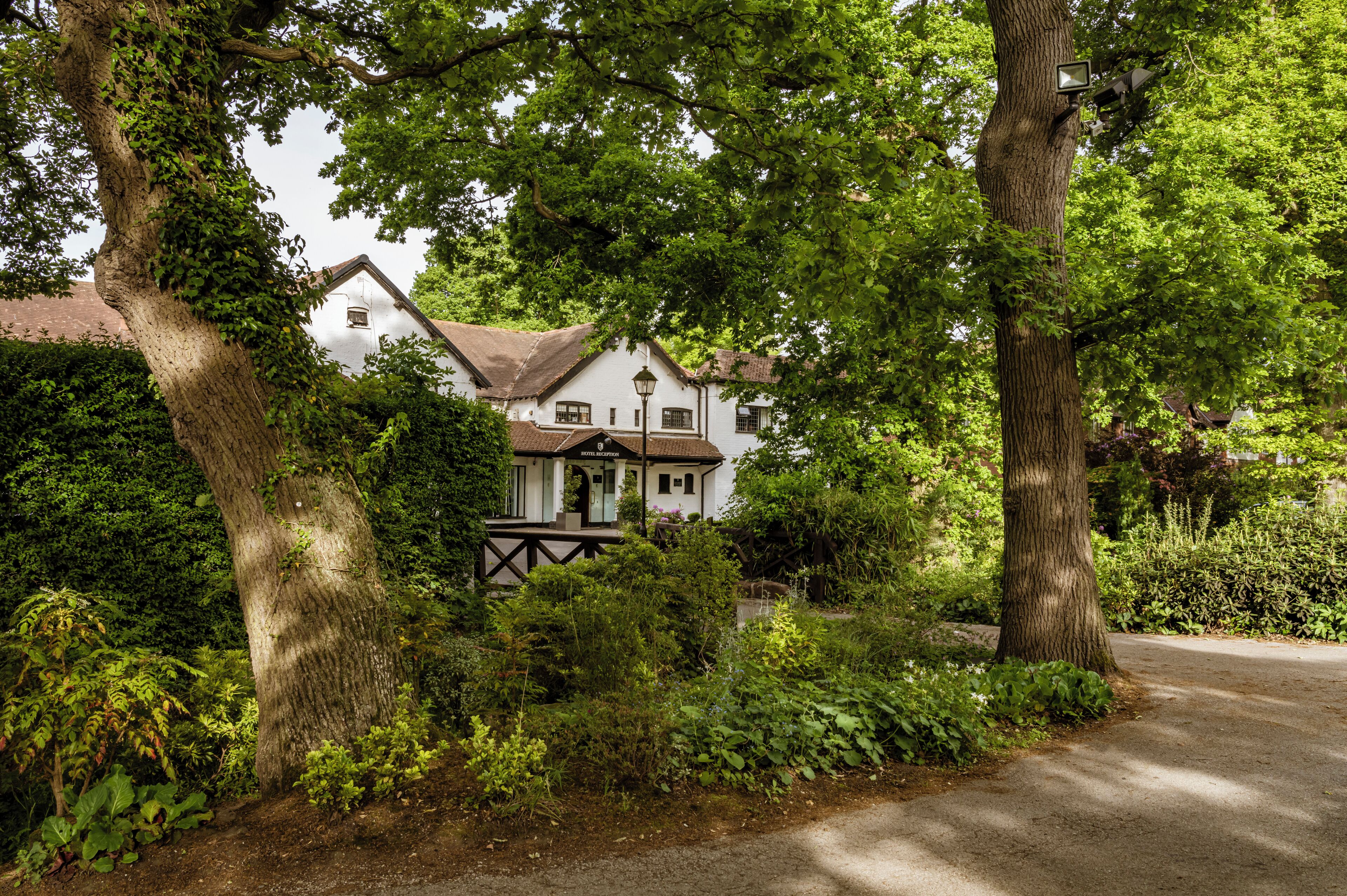 Un bâtiment traditionnel de couleur blanche, doté d’un porche à ossature en bois sombre, entouré d’une végétation luxuriante et d’arbres.