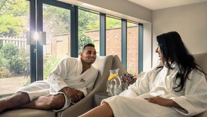 A couple in white bathrobes sitting on a beige sofa in a well-lit room with large windows, a small table with a vase of flowers, and a glass of orange juice. -  Macdonald Craxton Wood Hotel & Spa, Chester (Ellesmere Port)