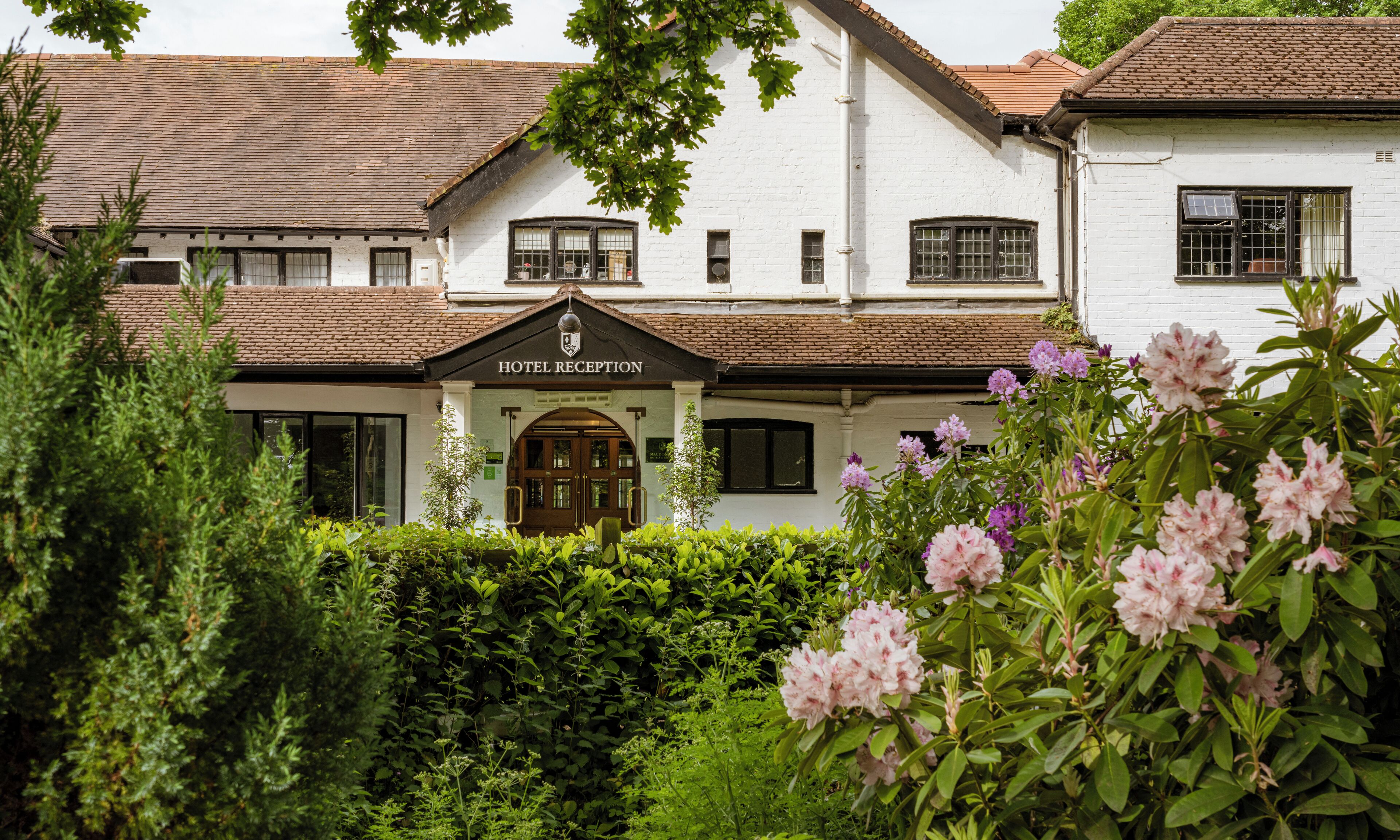 A white building with a brown roof, a sign reading "Hotel Reception", and a well-maintained garden with pink flowers.