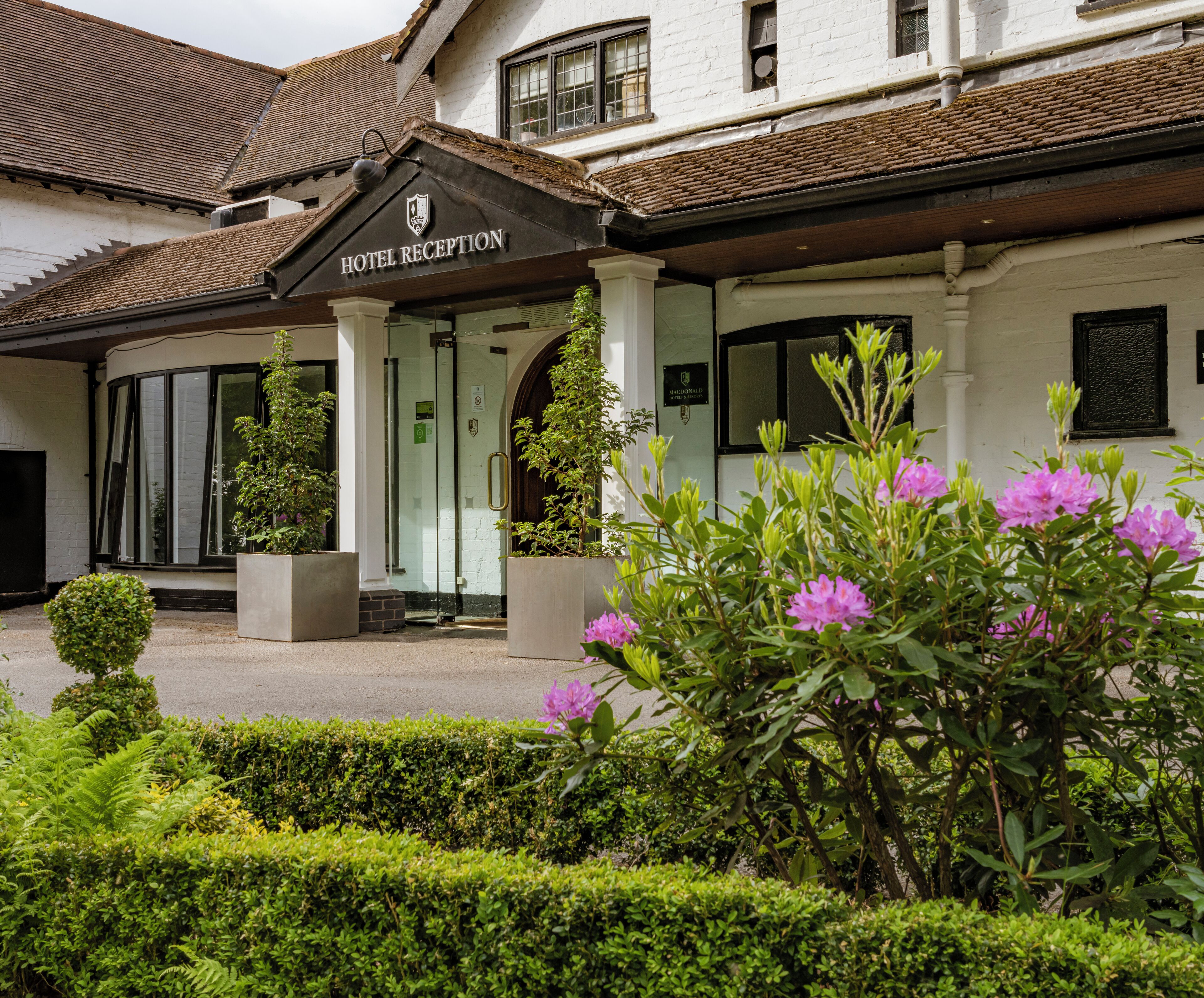 A hotel reception with a traditional design, featuring a signboard, a glass door entrance, and a well-maintained garden with flowering plants.