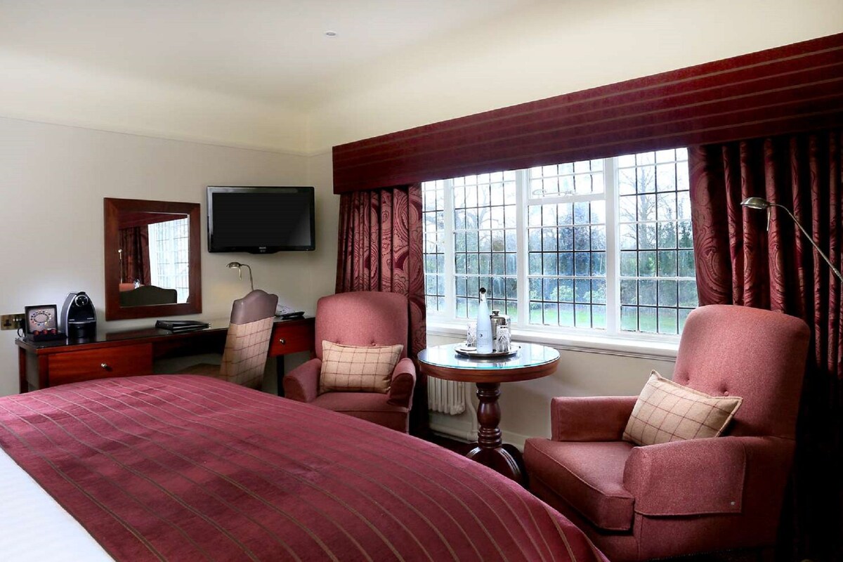 A hotel room with a large bed, two armchairs, a desk with a television, and a window with curtains.