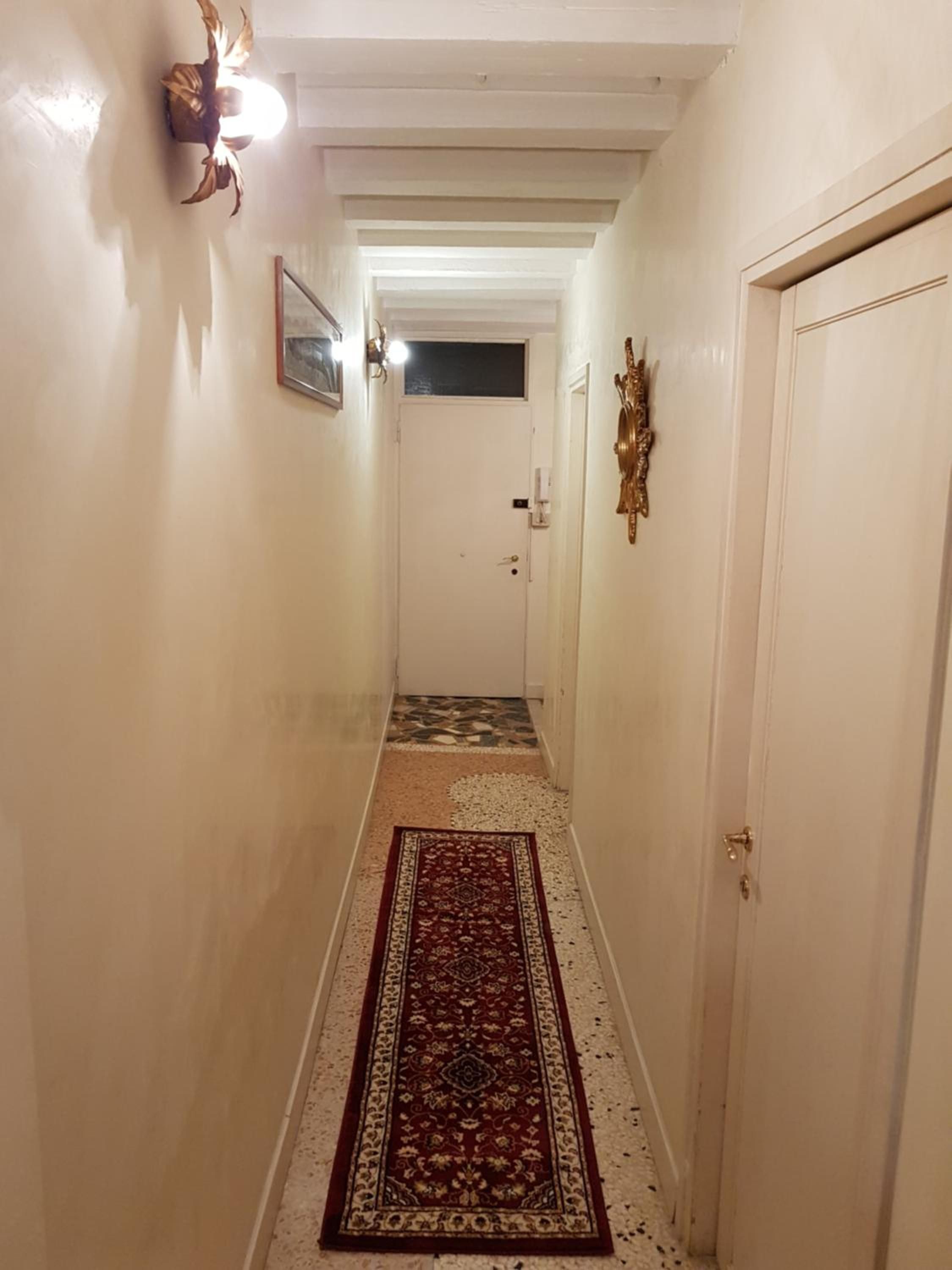 Un corridoio stretto con un tappeto decorato, una porta in fondo e lampade a parete.