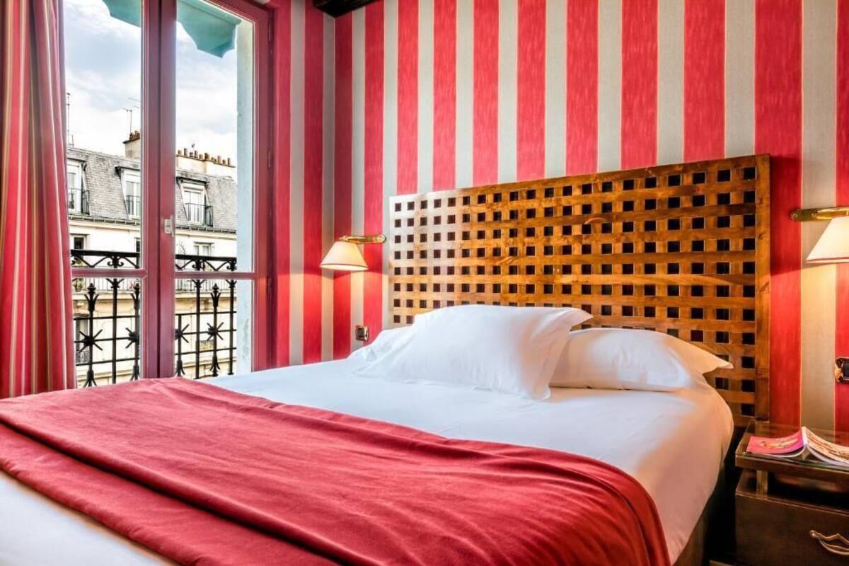 A hotel room with a striped red and white wall, a wooden headboard, a bed with white linens, and a view of the city through a window.