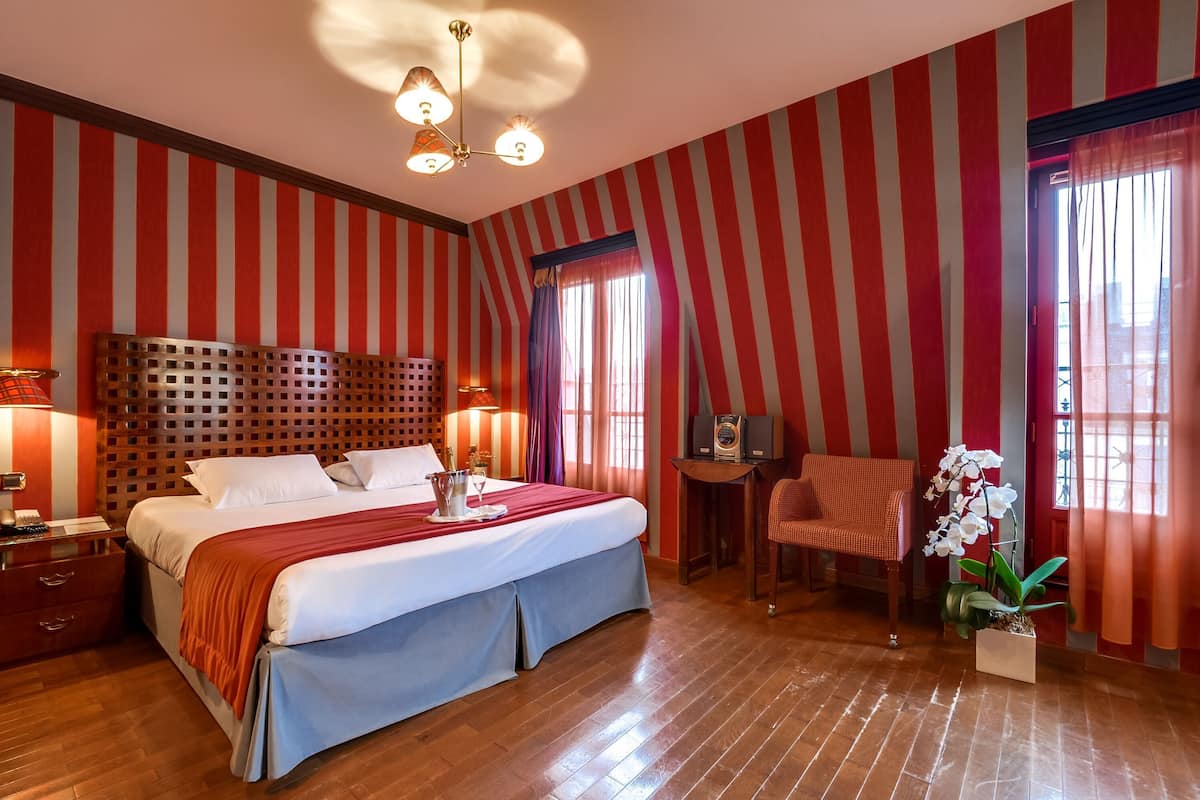 A hotel room with a large bed, a wooden headboard, a desk with a chair, a small table with a radio, and a potted orchid.