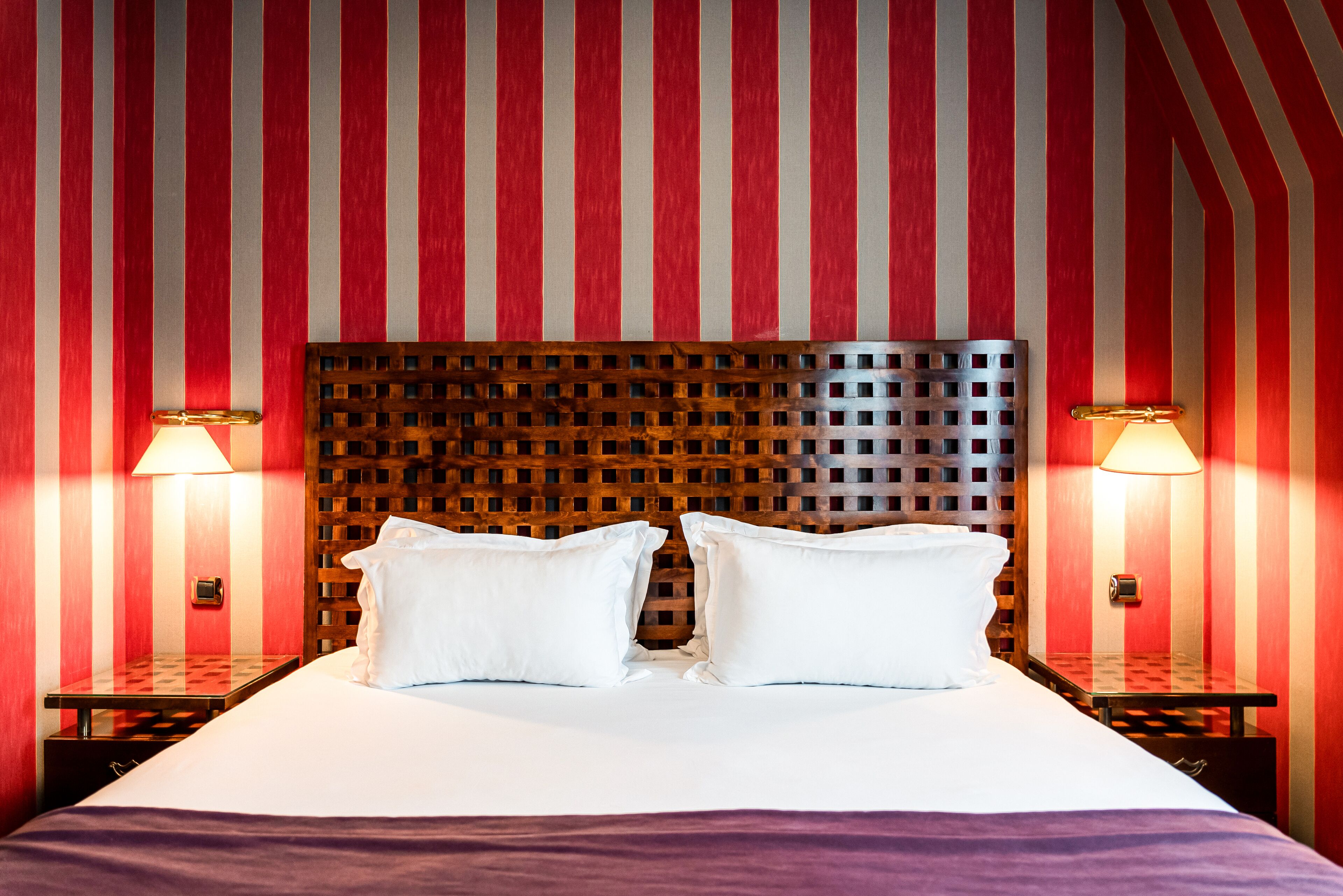 Habitación de hotel con pared a rayas rojas y blancas, cabecera de madera, dos mesitas de noche con lámparas y una cama bien tendida con sábanas blancas.