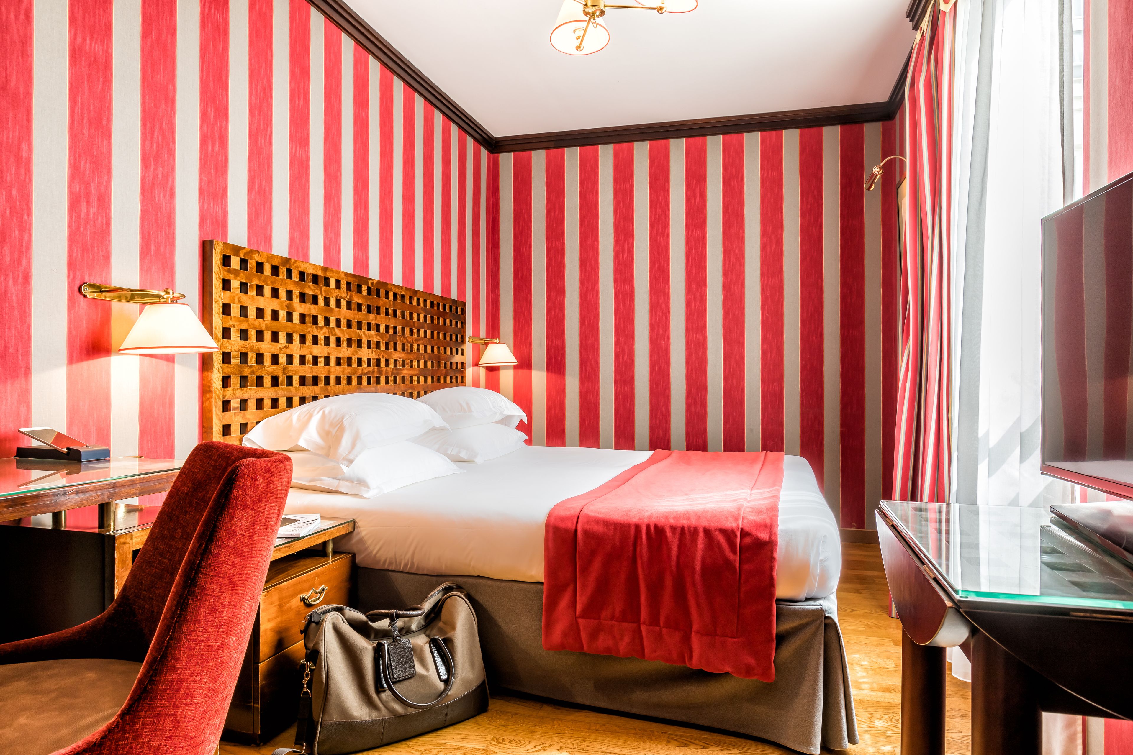 Habitación de hotel con pared a rayas rojas y blancas, cabecera de madera, cama con ropa blanca, manta roja, escritorio con lámpara, una silla y un televisor.