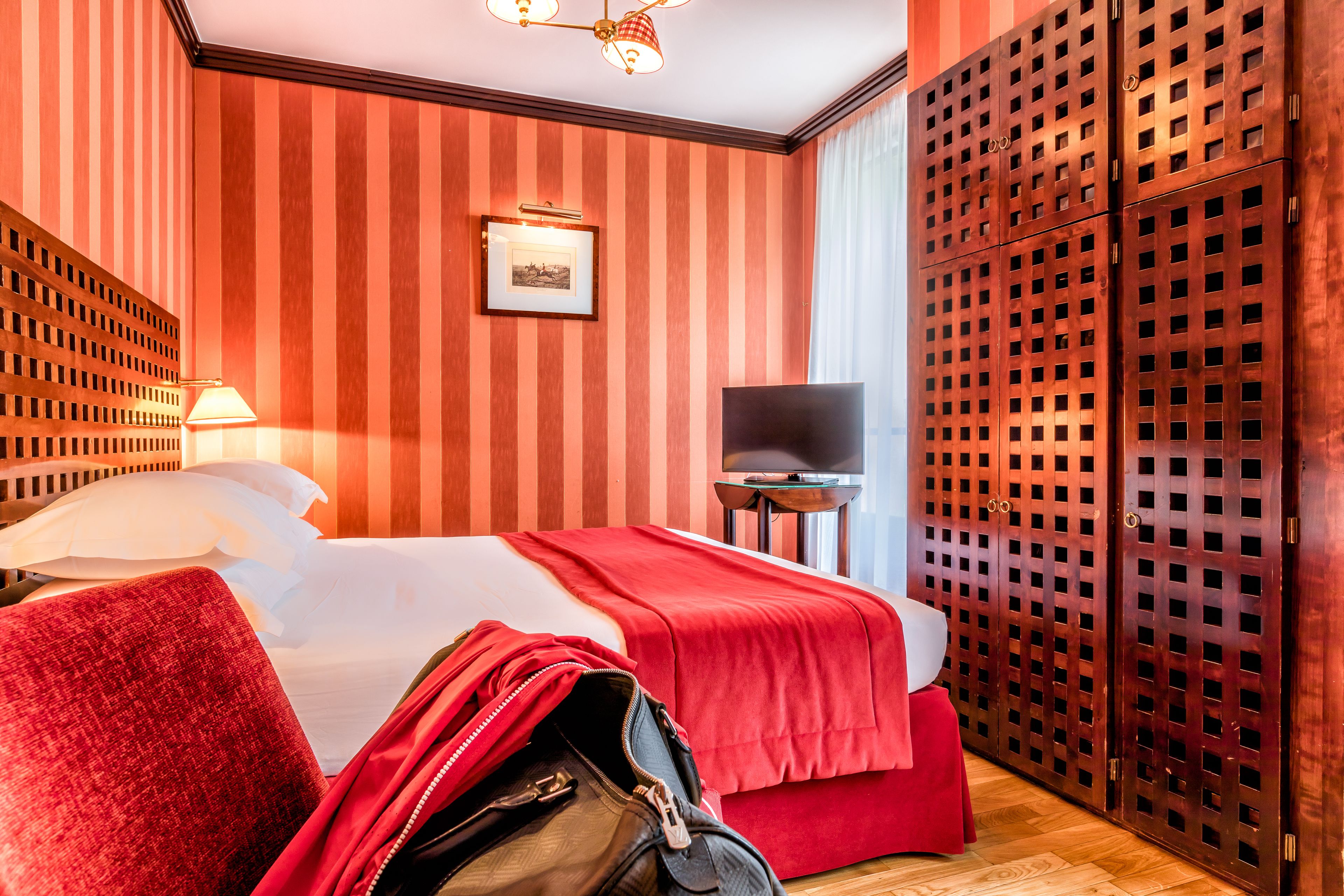 Quarto de hotel com cama, televisão, guarda-roupa de madeira e mala vermelha no chão.