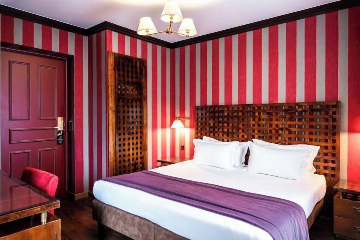 A hotel room with a striped wall, a wooden headboard, a bed with white linens, a desk with a chair, and a nightstand with a lamp.