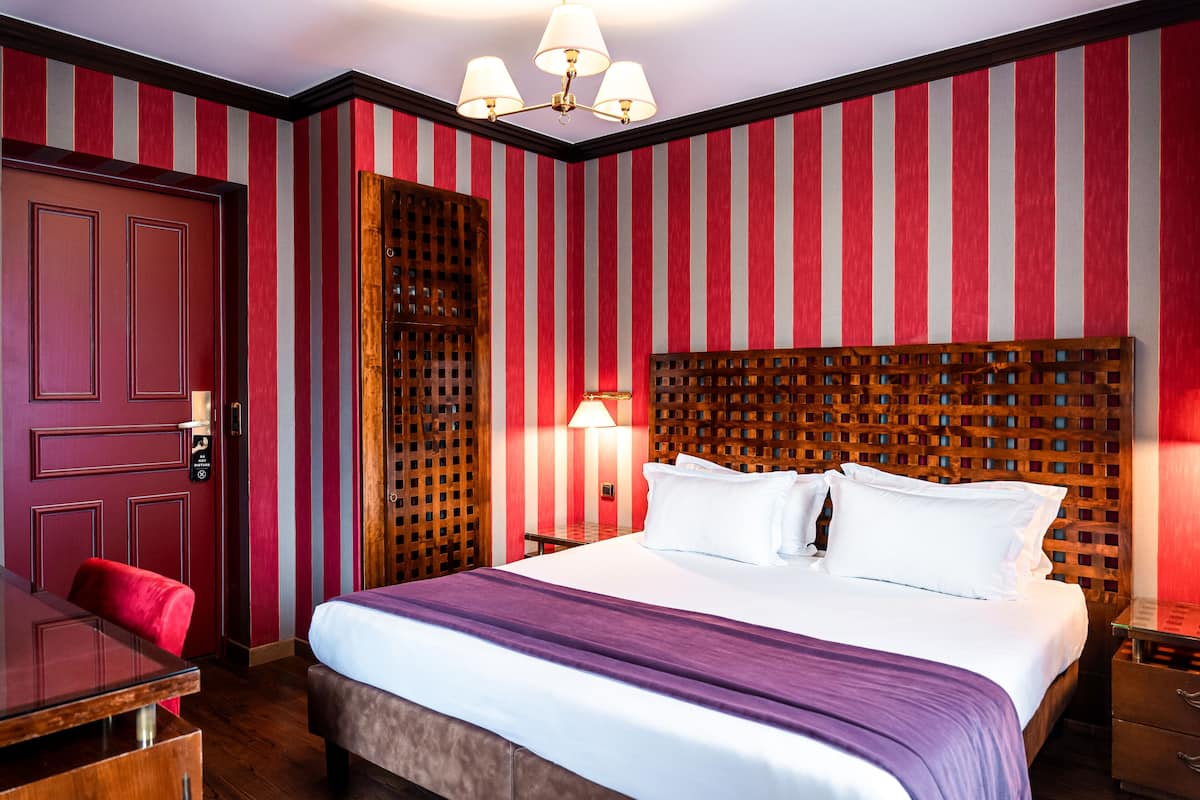 A hotel room with a large bed, a wooden headboard, a desk with a chair, and a red-striped wall.