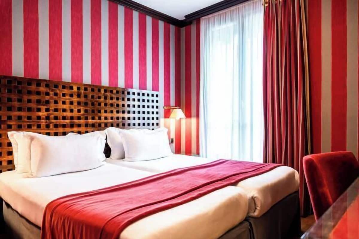A hotel room with a bed, a wooden headboard, a red and white striped wall, a window with curtains, and a red chair.