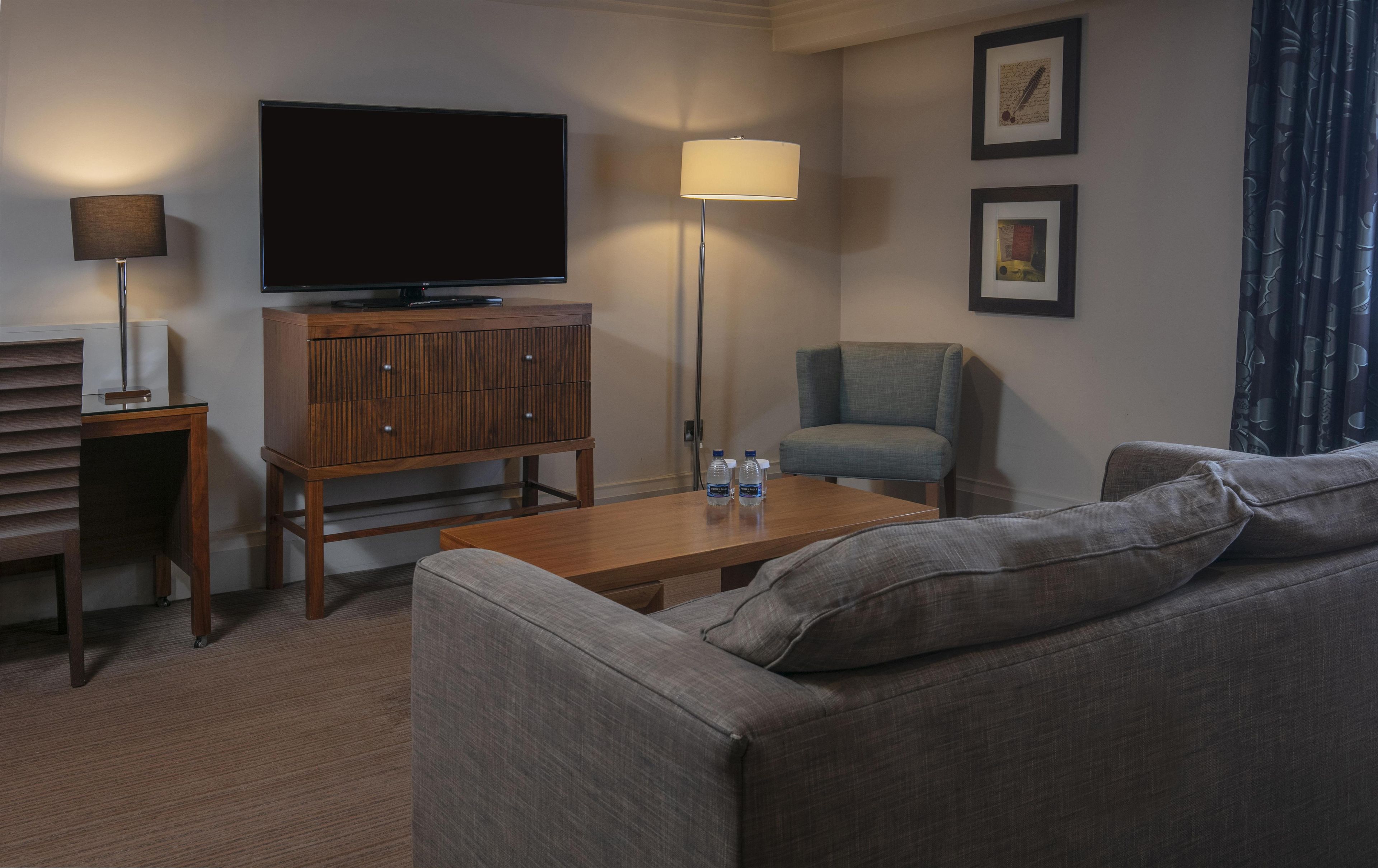 A hotel room with a flat-screen TV, a sofa, a chair, a coffee table with water bottles, and a lamp.