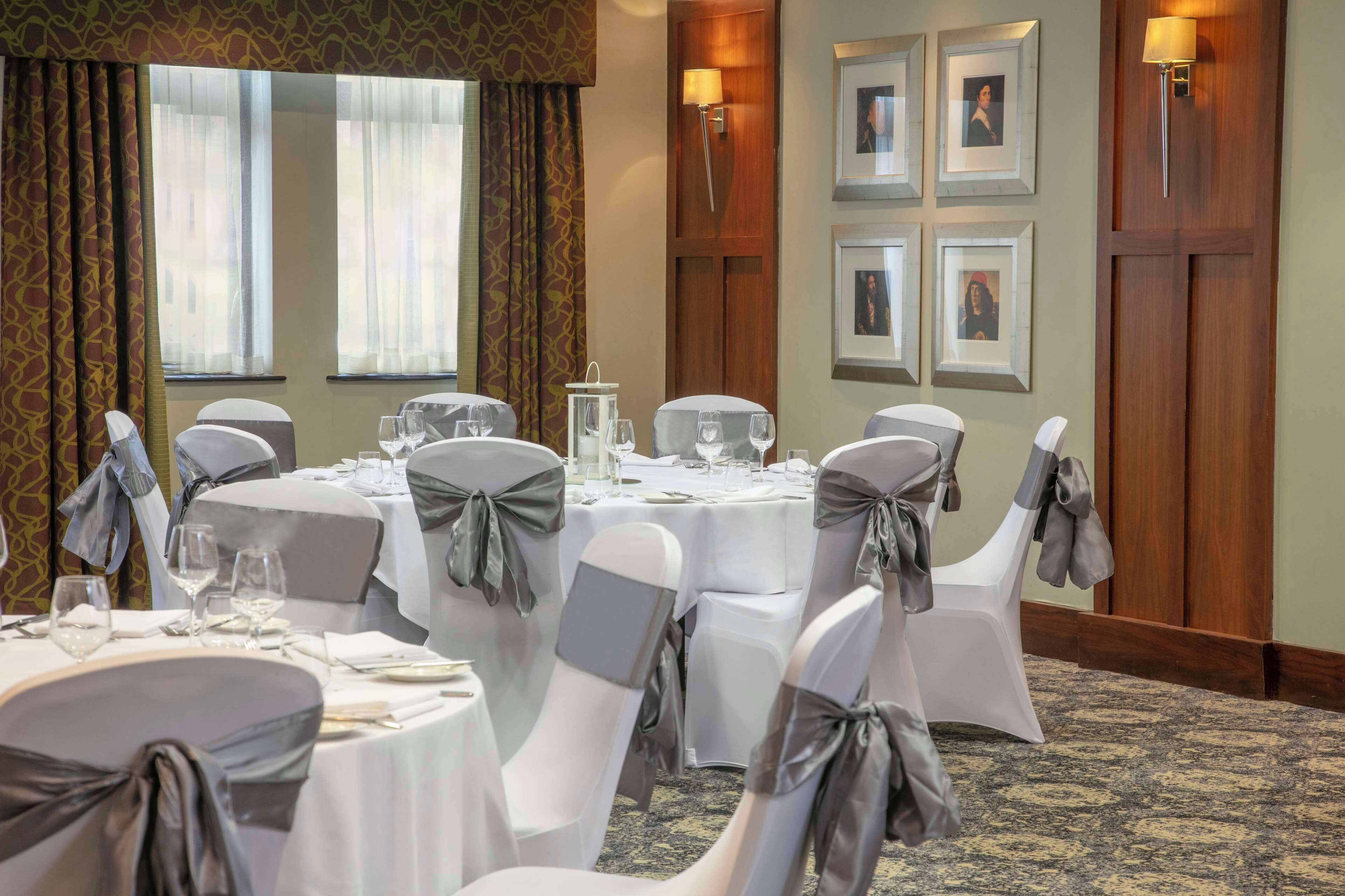 Uma sala de jantar de hotel com mesas redondas, cadeiras com fitas cinzas, quadros nas paredes e piso coberto de carpete.