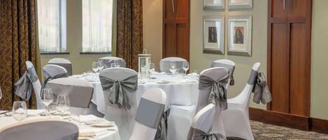 Uma sala de jantar de hotel com mesas redondas, cadeiras com fitas cinzas, quadros nas paredes e piso coberto de carpete.