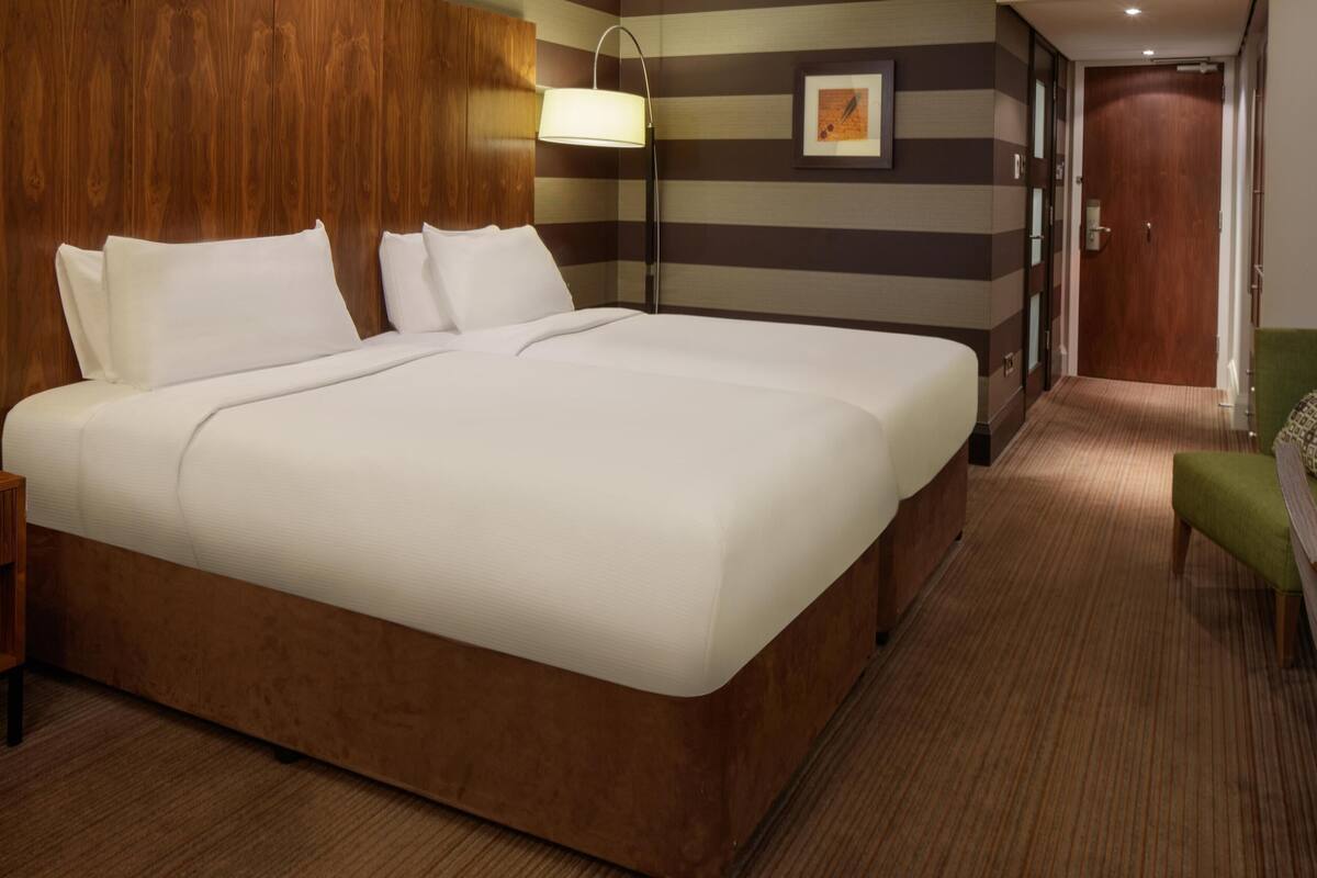 A hotel room with a large bed, a bedside lamp, a framed picture on the wall, and a green chair.