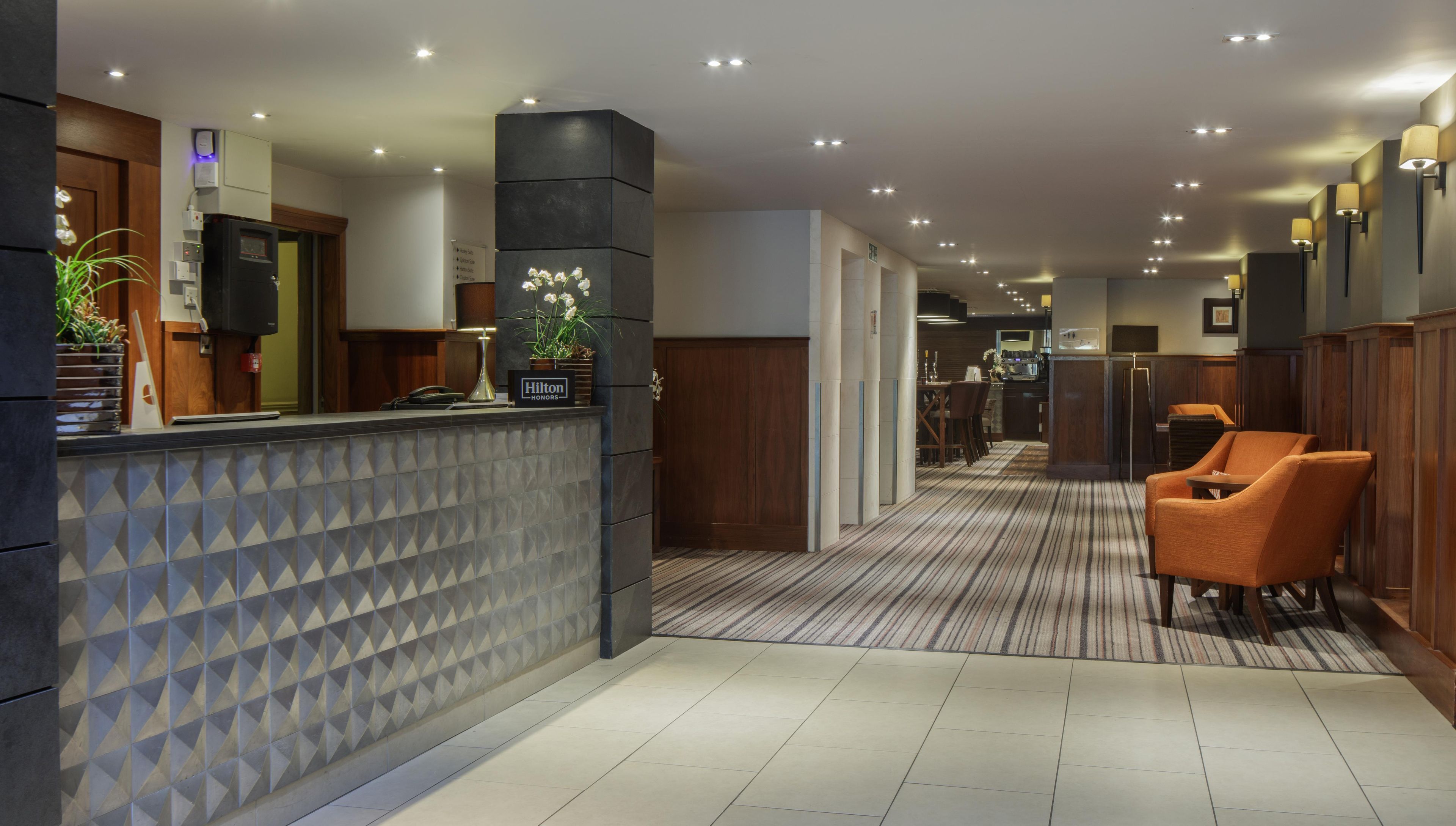A hotel reception area with a reception desk, a seating area with chairs, and a hallway leading to the dining area.