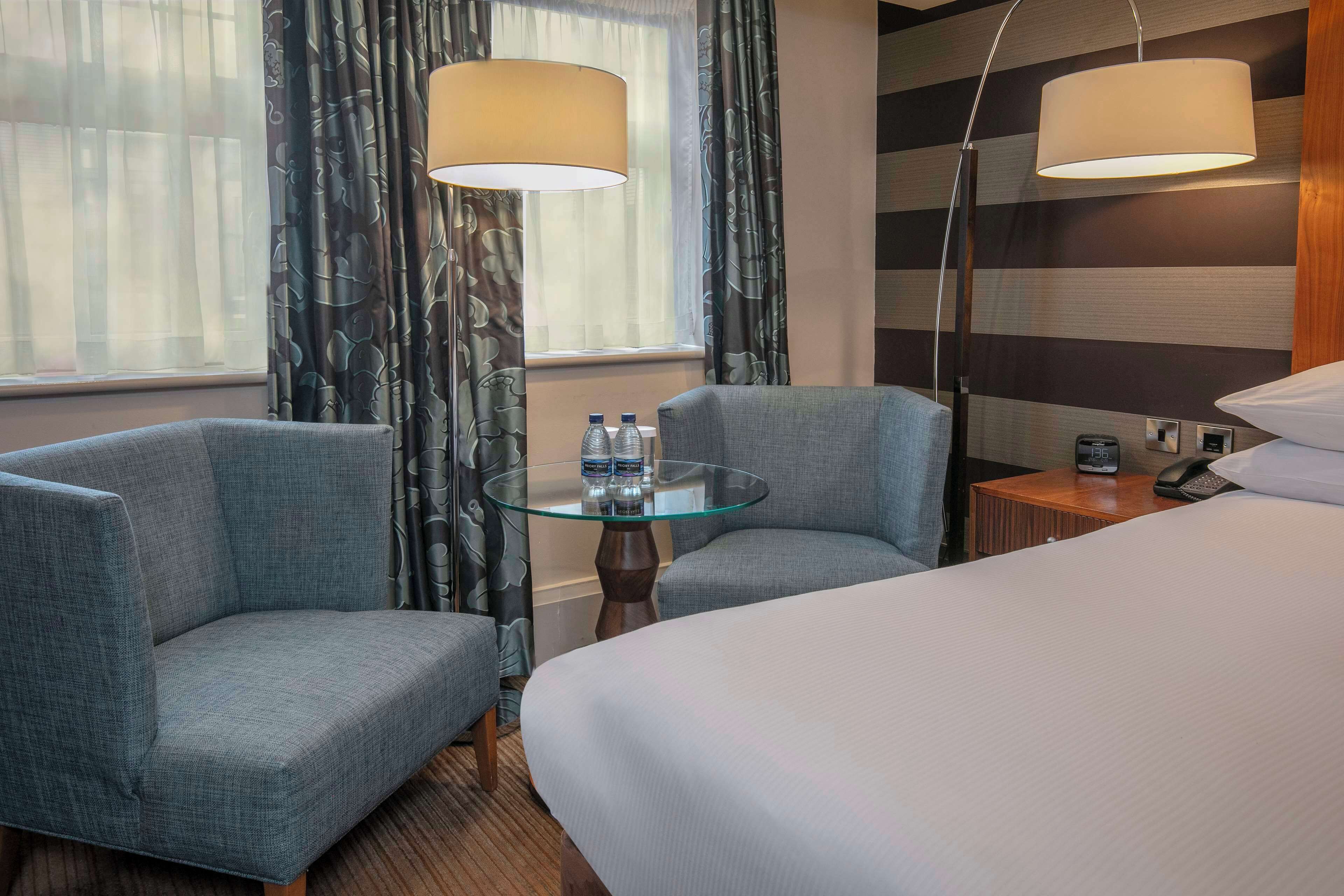Une chambre d’hôtel avec un lit, deux fauteuils, une table en verre avec des bouteilles d’eau, une lampe, une horloge et un téléphone sur une table de chevet en bois.