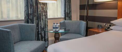 Une chambre d’hôtel avec un lit, deux fauteuils, une table en verre avec des bouteilles d’eau, une lampe, une horloge et un téléphone sur une table de chevet en bois.