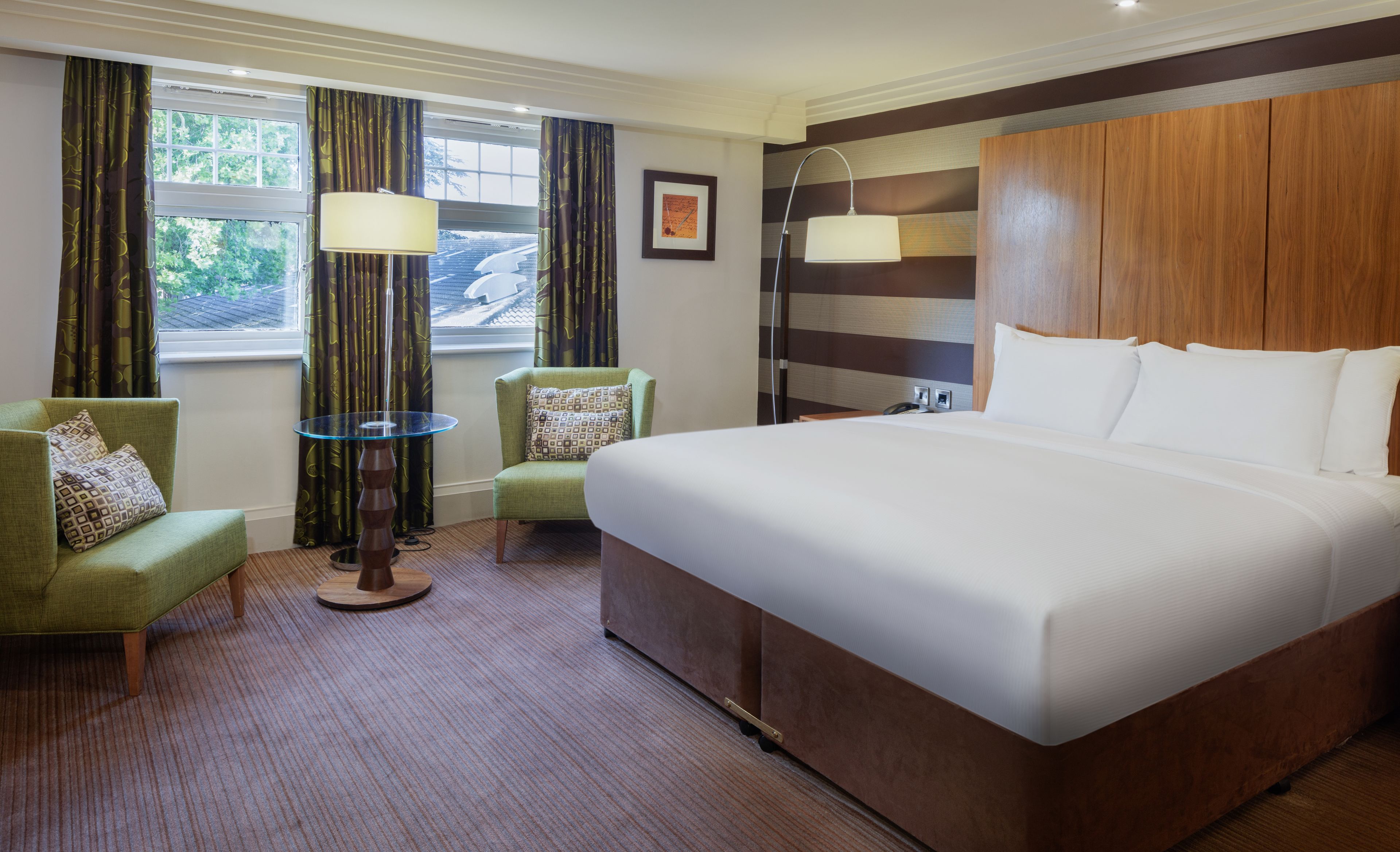 A hotel room with a large bed, a green armchair, a small table, and a window with curtains.