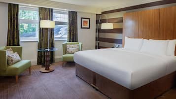 A hotel room with a large bed, a green armchair, a small table, and a window with curtains.