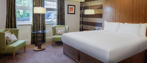 A hotel room with a large bed, a green armchair, a small table, and a window with curtains.