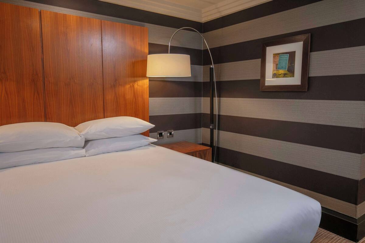 A hotel room with a large bed, a wooden headboard, a bedside table, a floor lamp, and a framed picture on the wall.