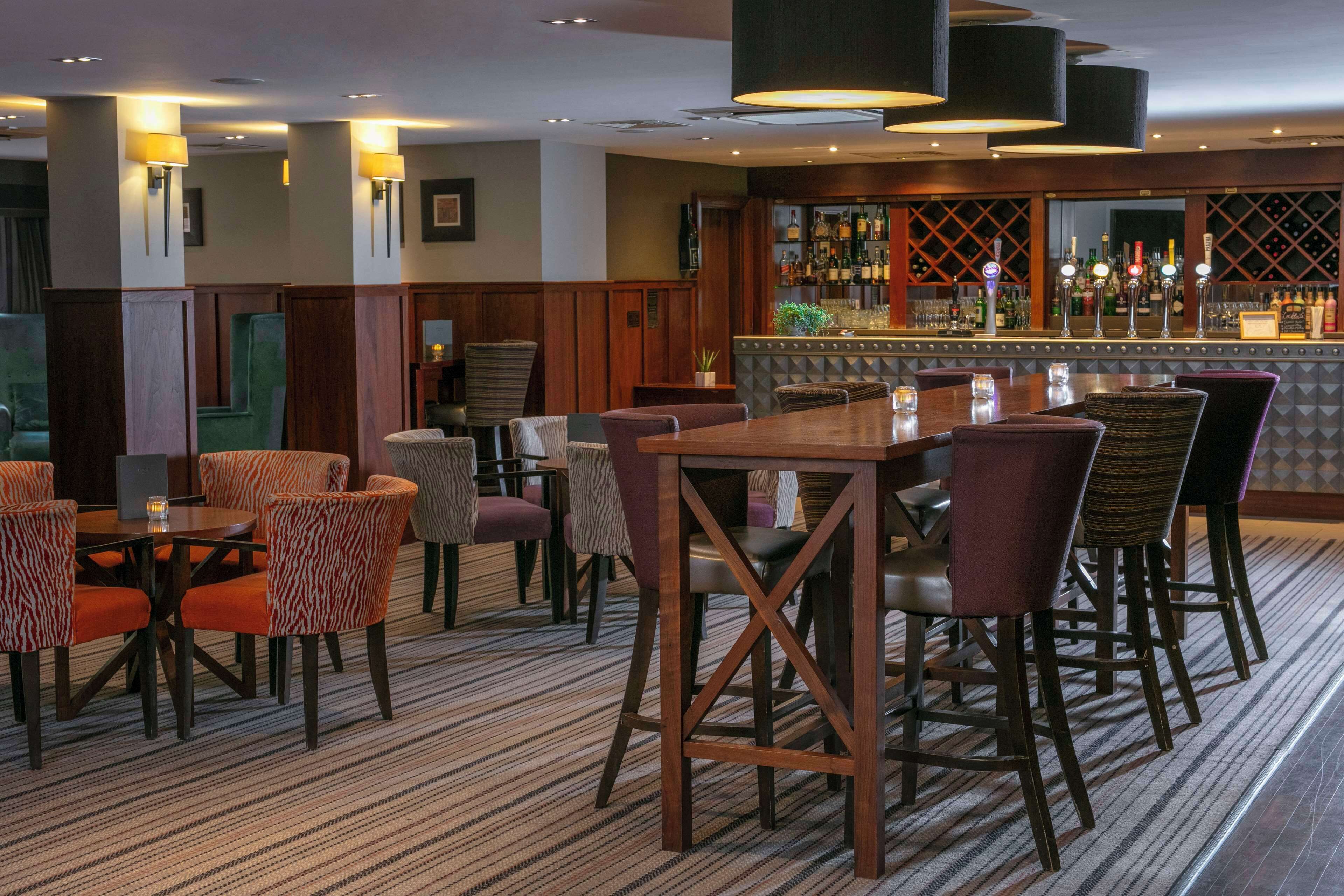 A hotel bar area with multiple seating arrangements, including round tables and bar stools. There are wall-mounted lamps and a bar counter with various bottles and glasses.