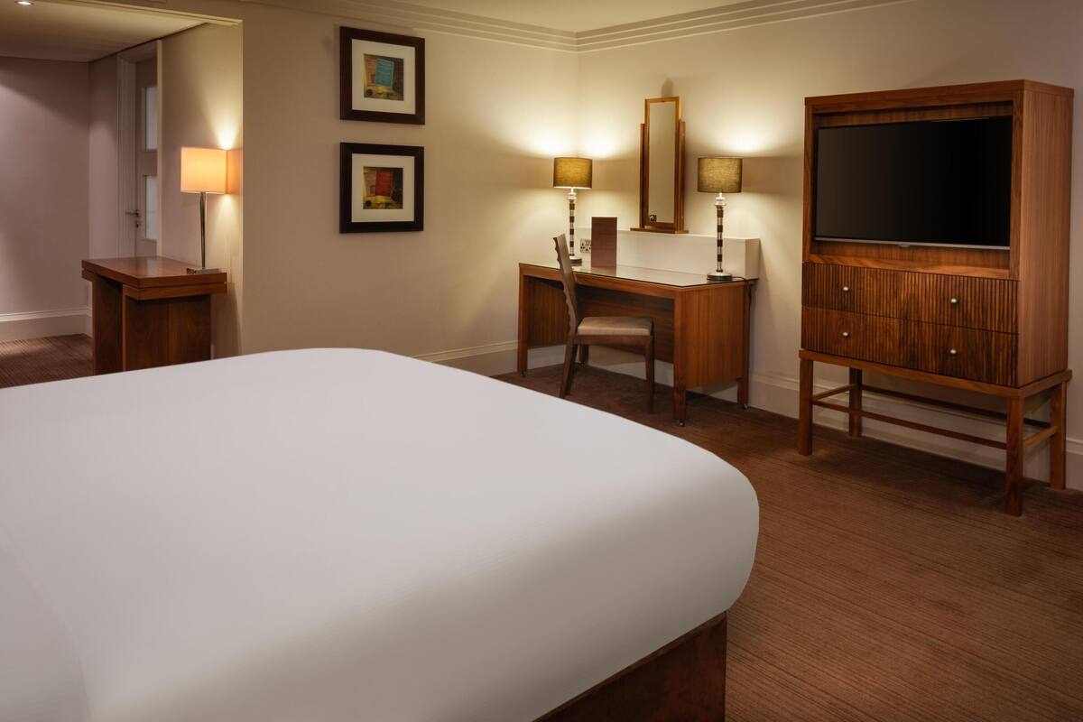 A hotel room with a large bed, a desk with a chair, a television, and framed pictures on the walls.