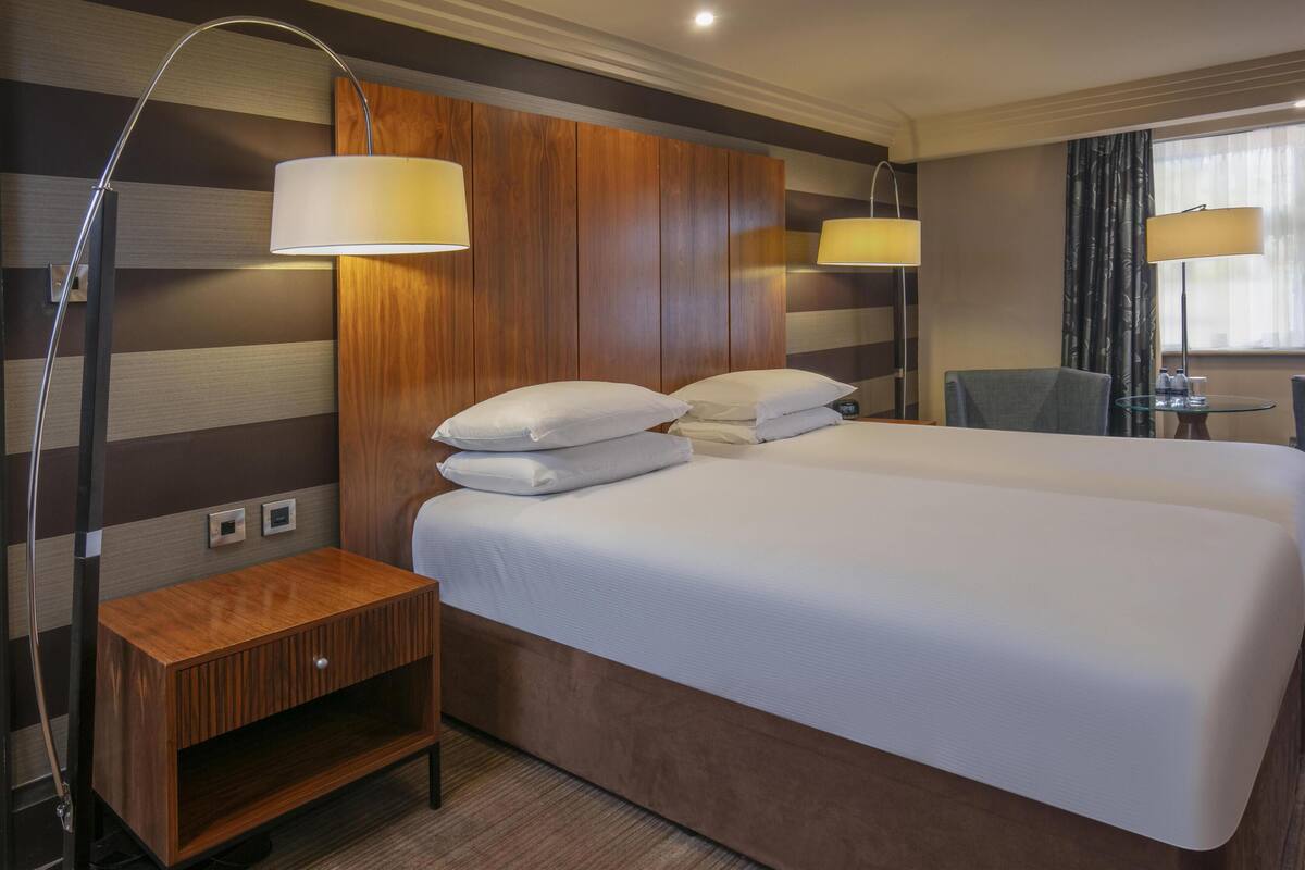 A hotel room with a large bed, two bedside lamps, a wooden nightstand, and a small table with a chair.