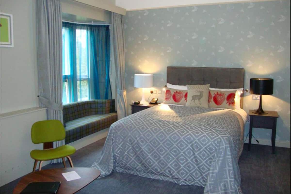 A bedroom with a bed, a nightstand, a lamp, a chair, and a window with curtains.