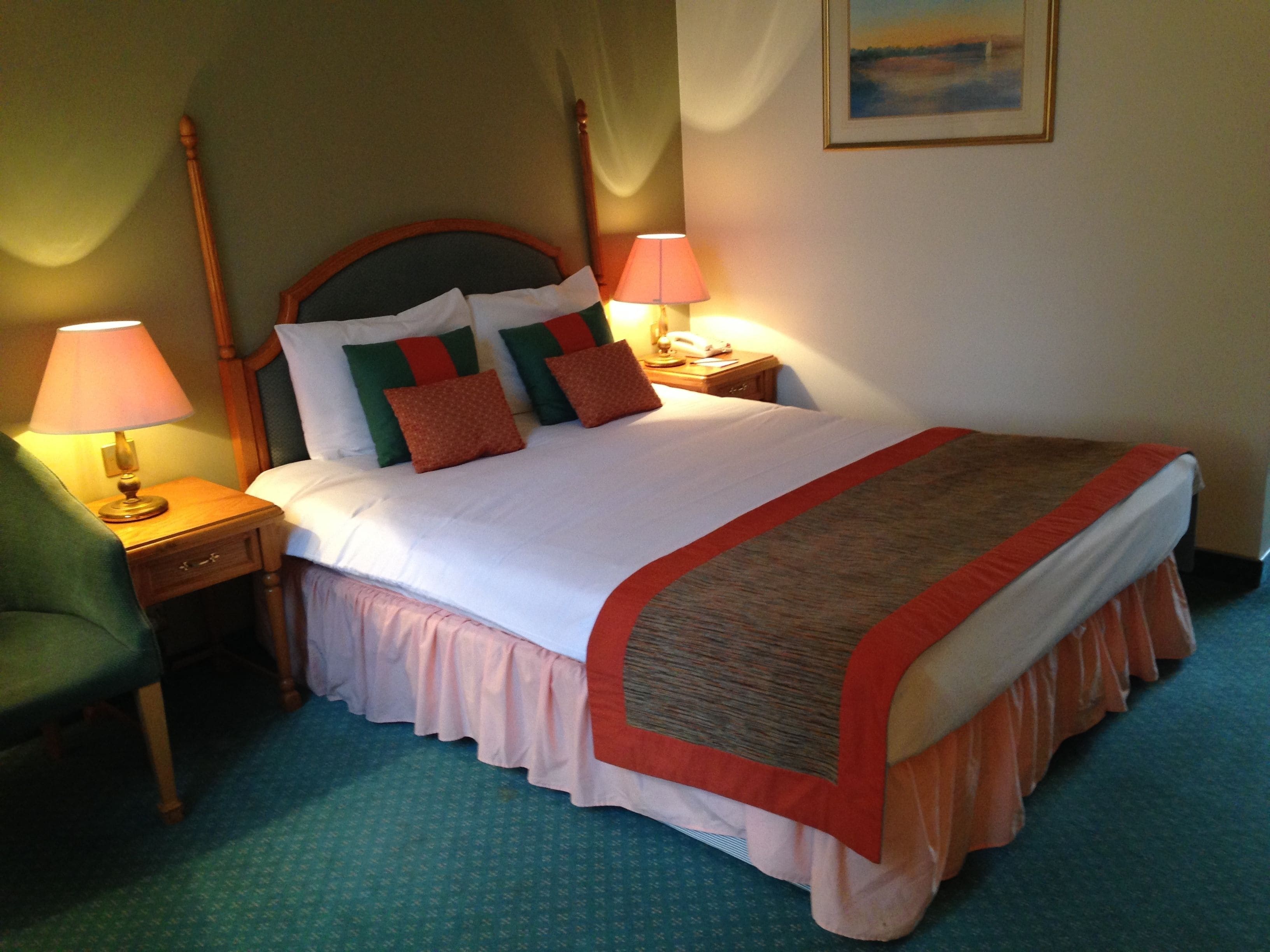 A neatly made bed with a white duvet, green and red pillows, and a brown throw. There are two bedside lamps on wooden nightstands.