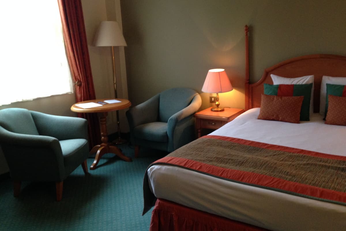 A hotel room with a bed, two armchairs, a small table, a lamp, and a window with curtains.