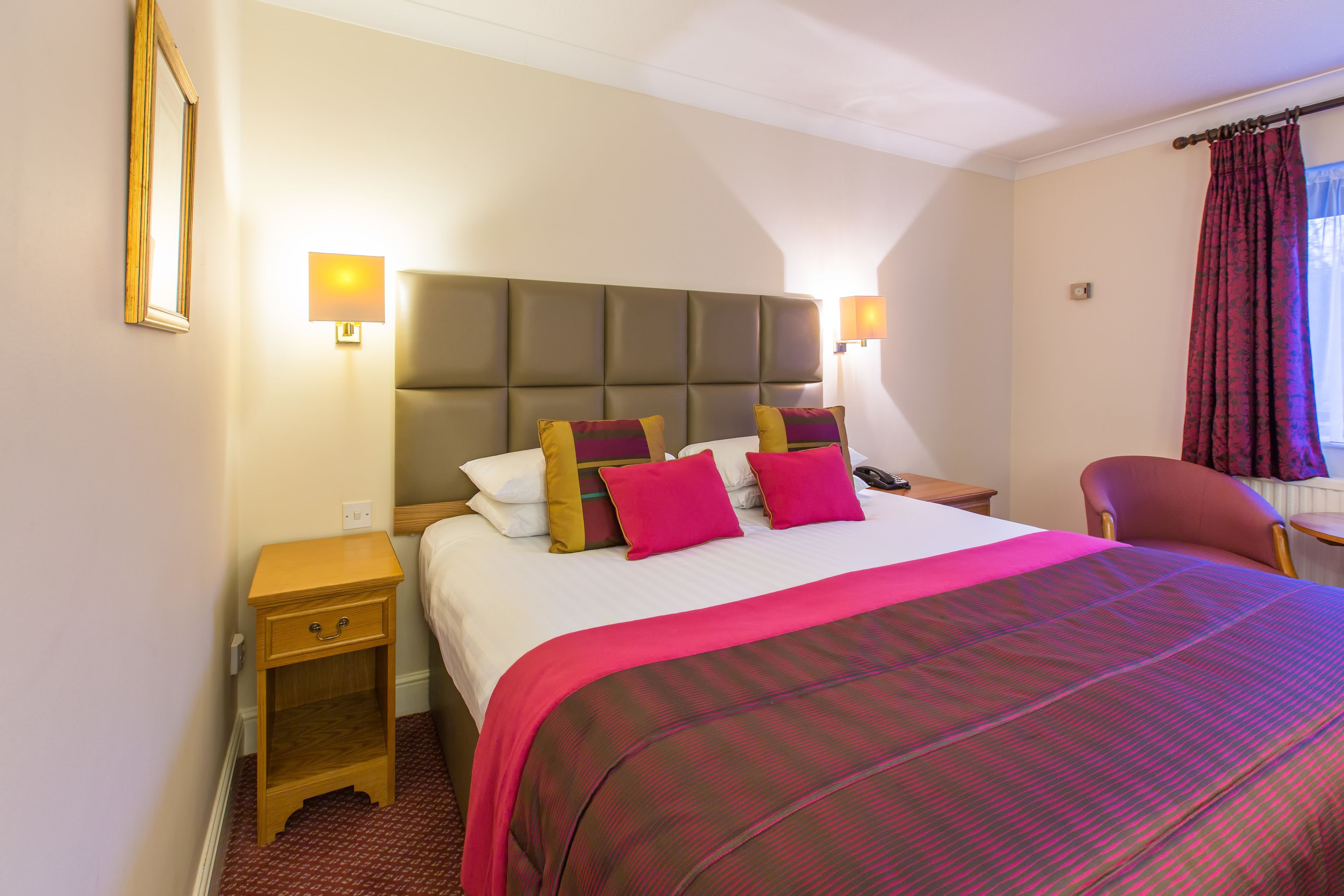 A hotel room with a large bed, two bedside lamps, a wooden nightstand, a chair, and a window with curtains.
