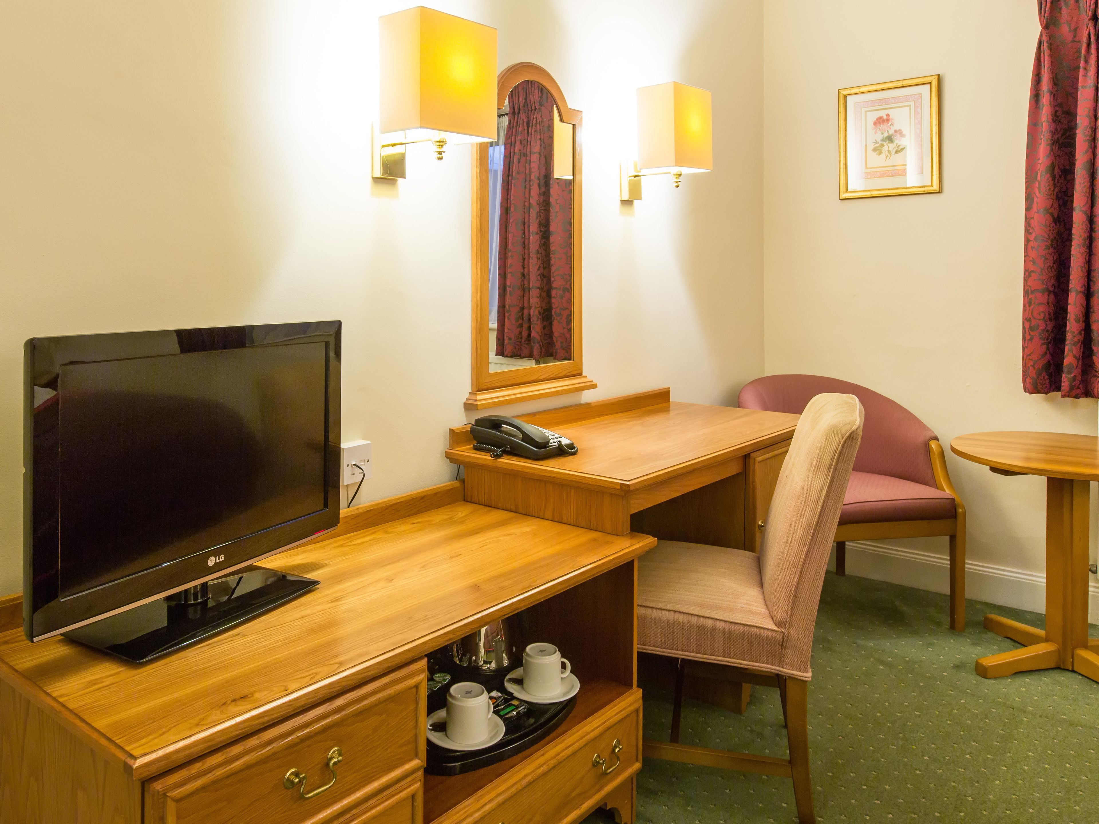 Ein Hotelzimmer mit einem Holztisch, einem Fernseher, einem Telefon, einem Sessel, einem kleinen Tisch und einem Spiegel.