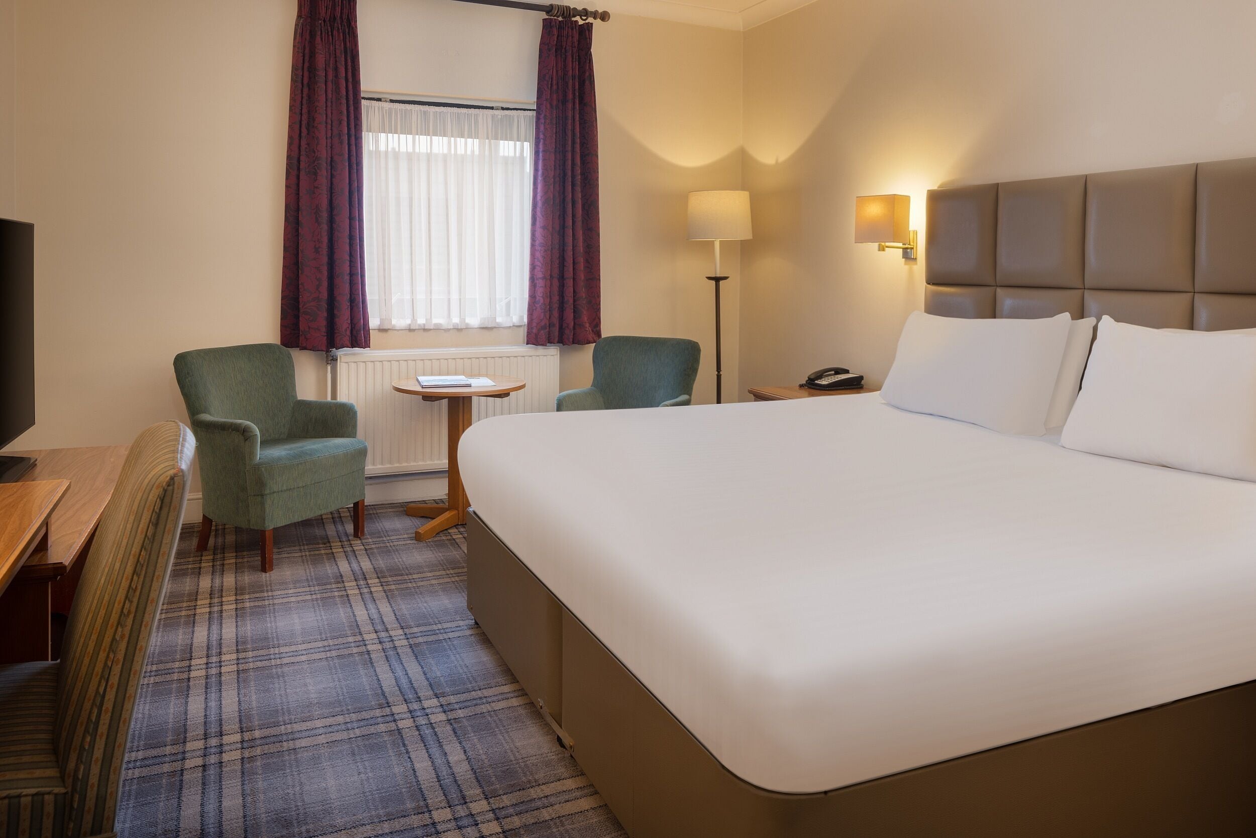A hotel room with a large bed, two armchairs, a small table, a lamp, and a television.