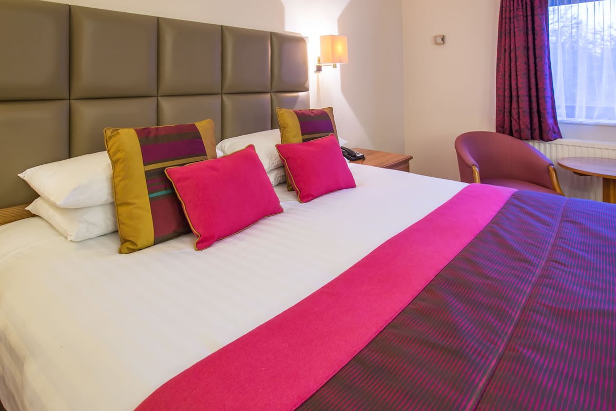 A hotel room with a large bed, multiple pillows, a nightstand, a chair, and a window with curtains.