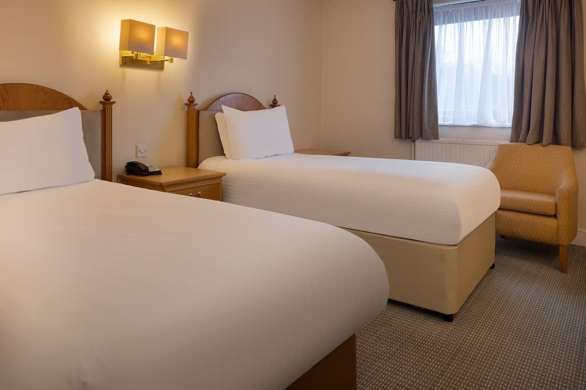 A hotel room with two beds, a chair, a nightstand with a phone, and wall-mounted lamps.