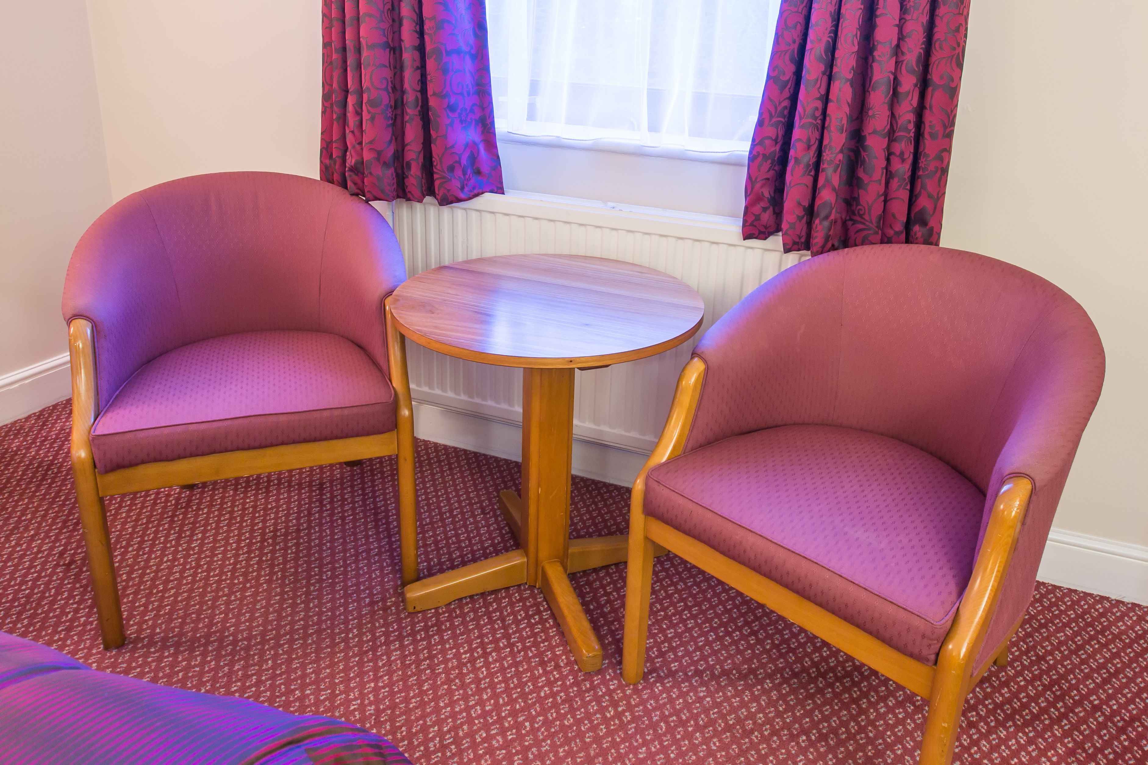 Two armchairs and a small round table in a room with patterned carpet and a window with curtains.
