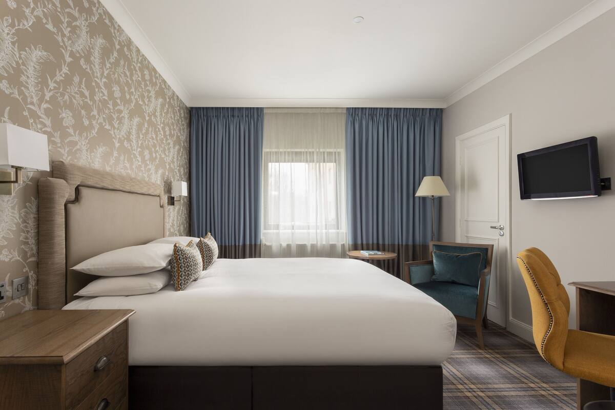 A hotel room with a bed, a desk, a chair, a TV, and a window with curtains.