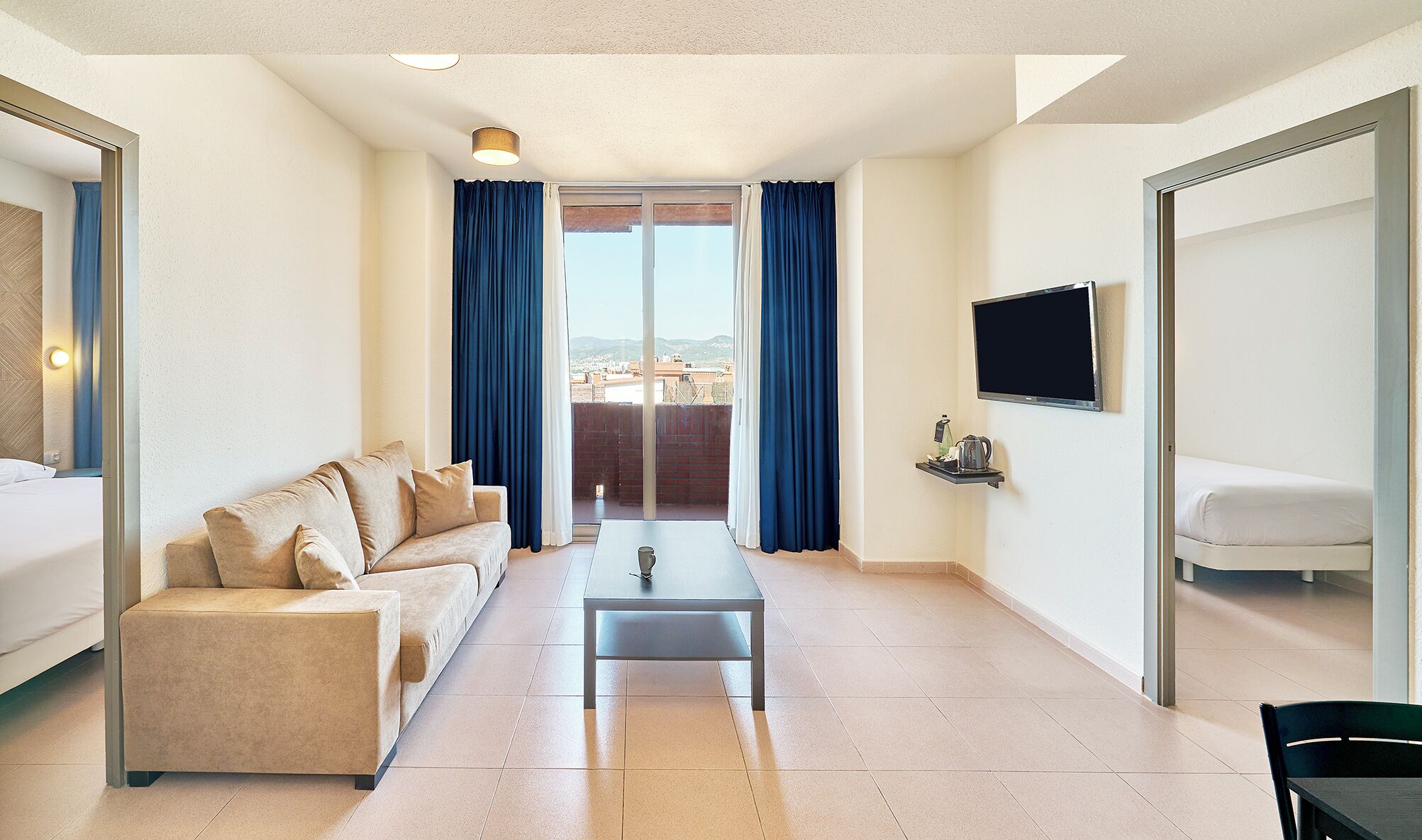 A hotel room with a sofa, a coffee table, a TV mounted on the wall, and a balcony with blue curtains.