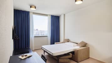 Habitación de hotel con cama, escritorio y silla. Tiene una ventana con cortinas, un televisor de pantalla plana montado en la pared y una lámpara en el techo.