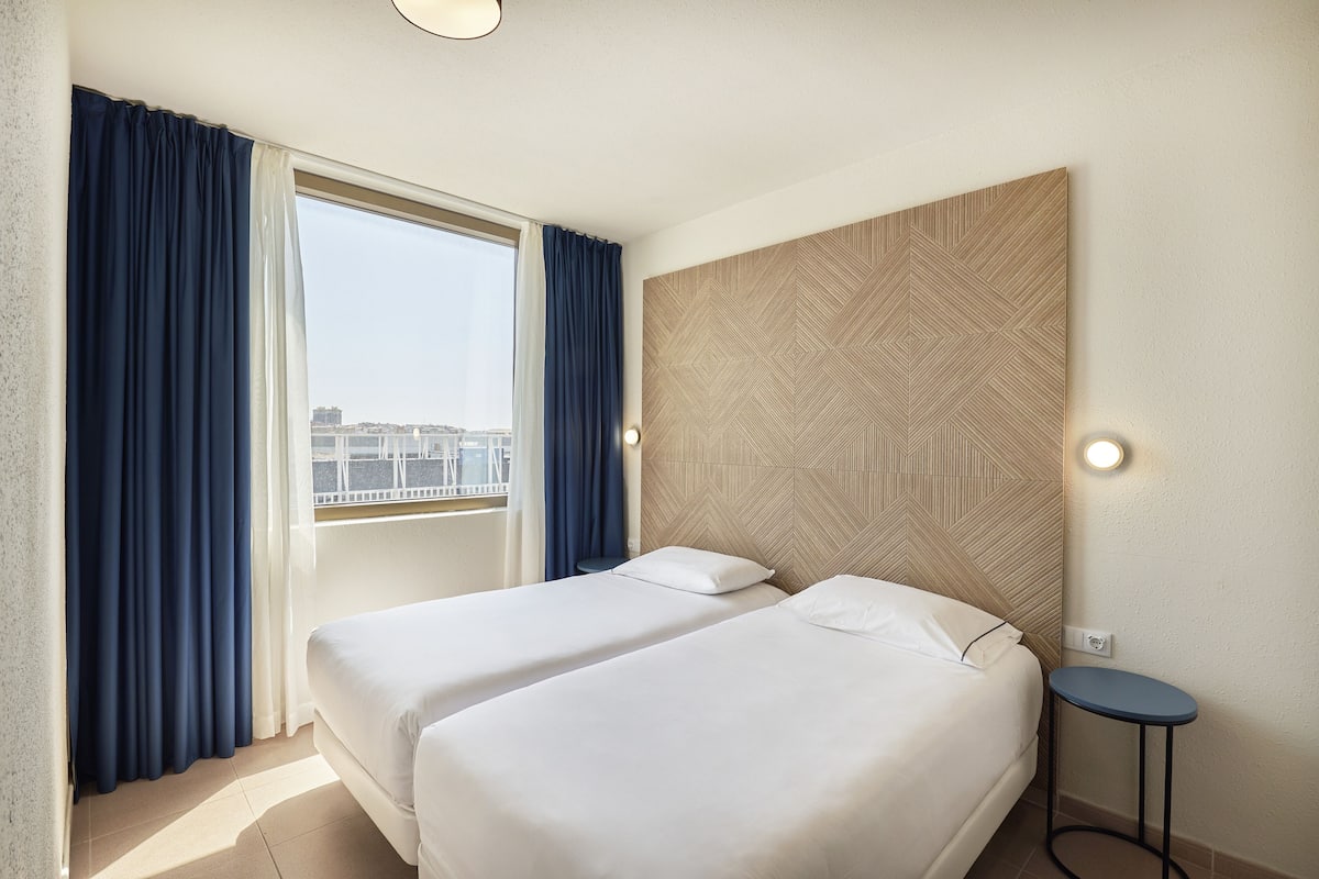 A hotel room with two beds, a large window with curtains, a wall-mounted light, and a small round table.