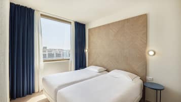 Habitación de hotel con dos camas, un ventanal con cortinas, una lámpara de pared y una pequeña mesa redonda.