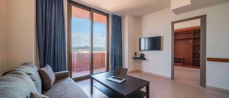 Una sala de estar moderna con un sofá, una mesa de centro y una televisión. Hay un ventanal con cortinas, un balcón y vistas de la ciudad.