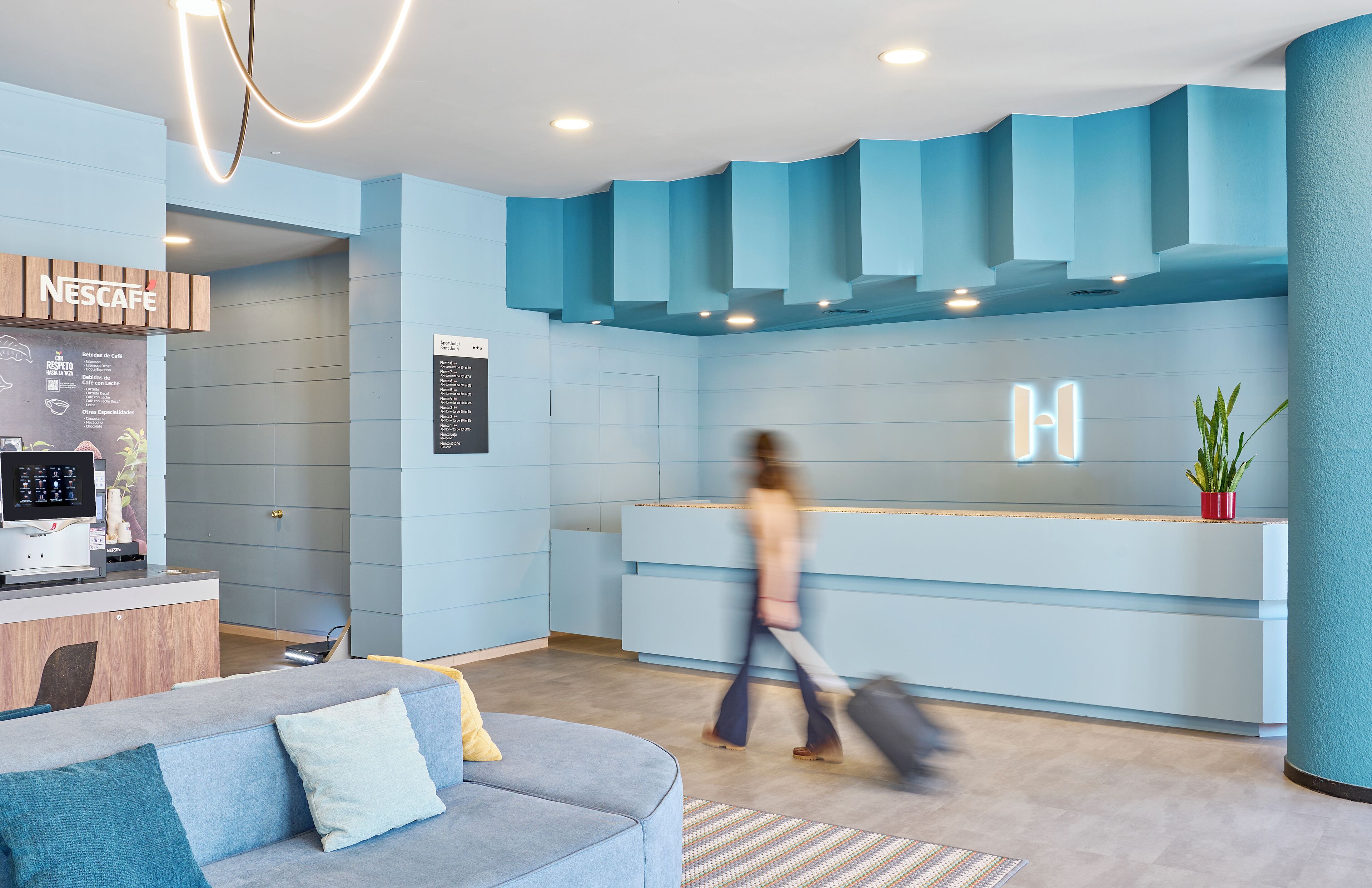 Un área de recepción moderna con una decoración azul, una máquina de café Nescafé y un espacio de estar con un sofá y cojines.