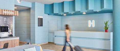 Un área de recepción moderna con una decoración azul, una máquina de café Nescafé y un espacio de estar con un sofá y cojines.
