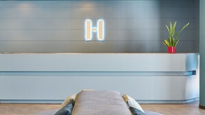 A modern hotel lobby with a grey bench, a neon 'H' sign, and a potted plant. - Apartamentos Hesperia Sant Joan (Sant Joan Despi)