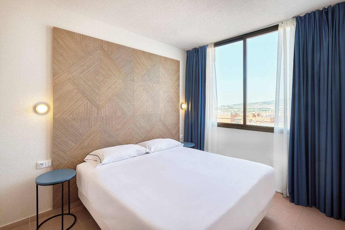 A hotel room with a large bed, a small side table, a wall-mounted light, and a window with curtains.