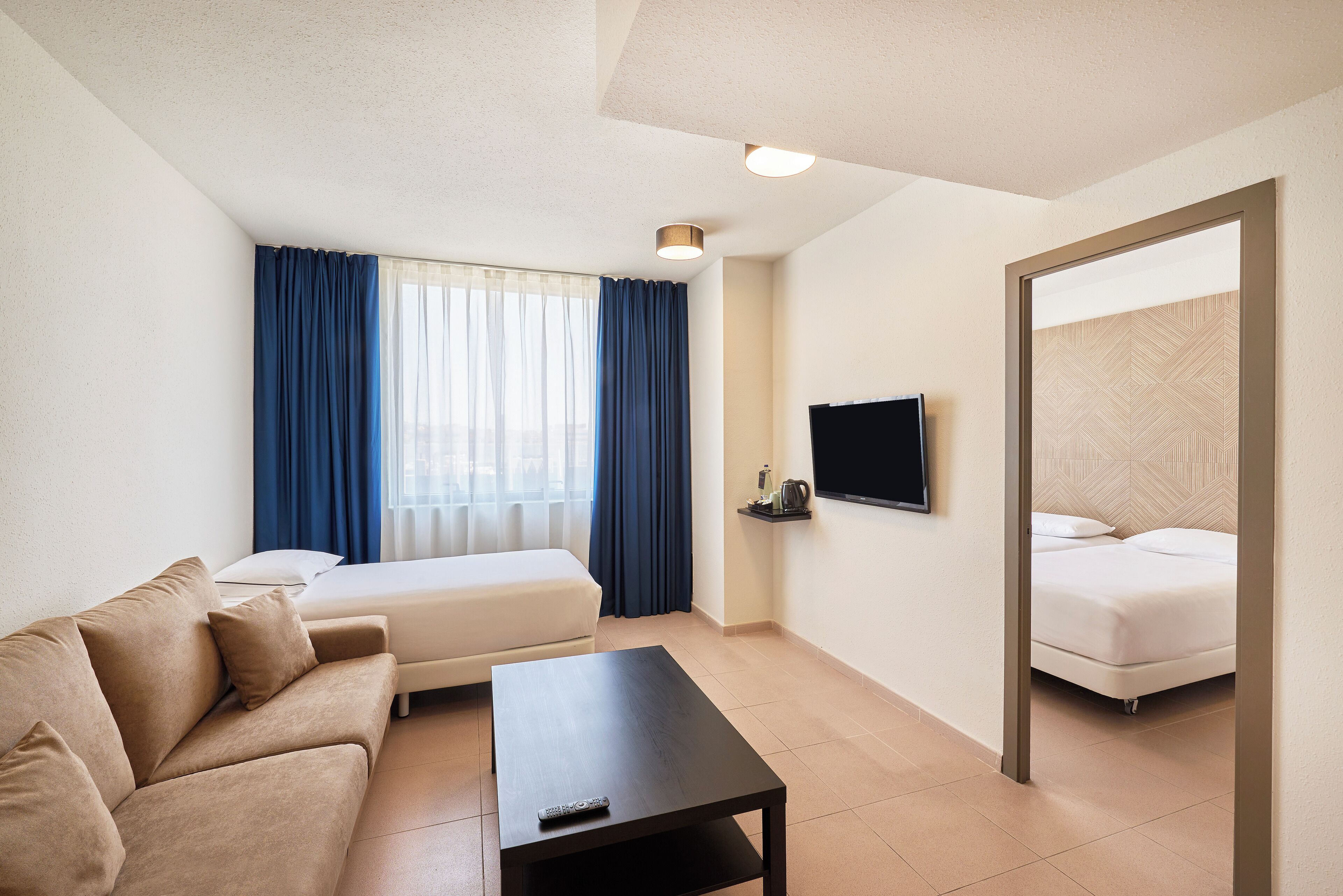 Et hotelværelse med sofa, lille bord, seng, fjernsyn og et vindue med gardiner.