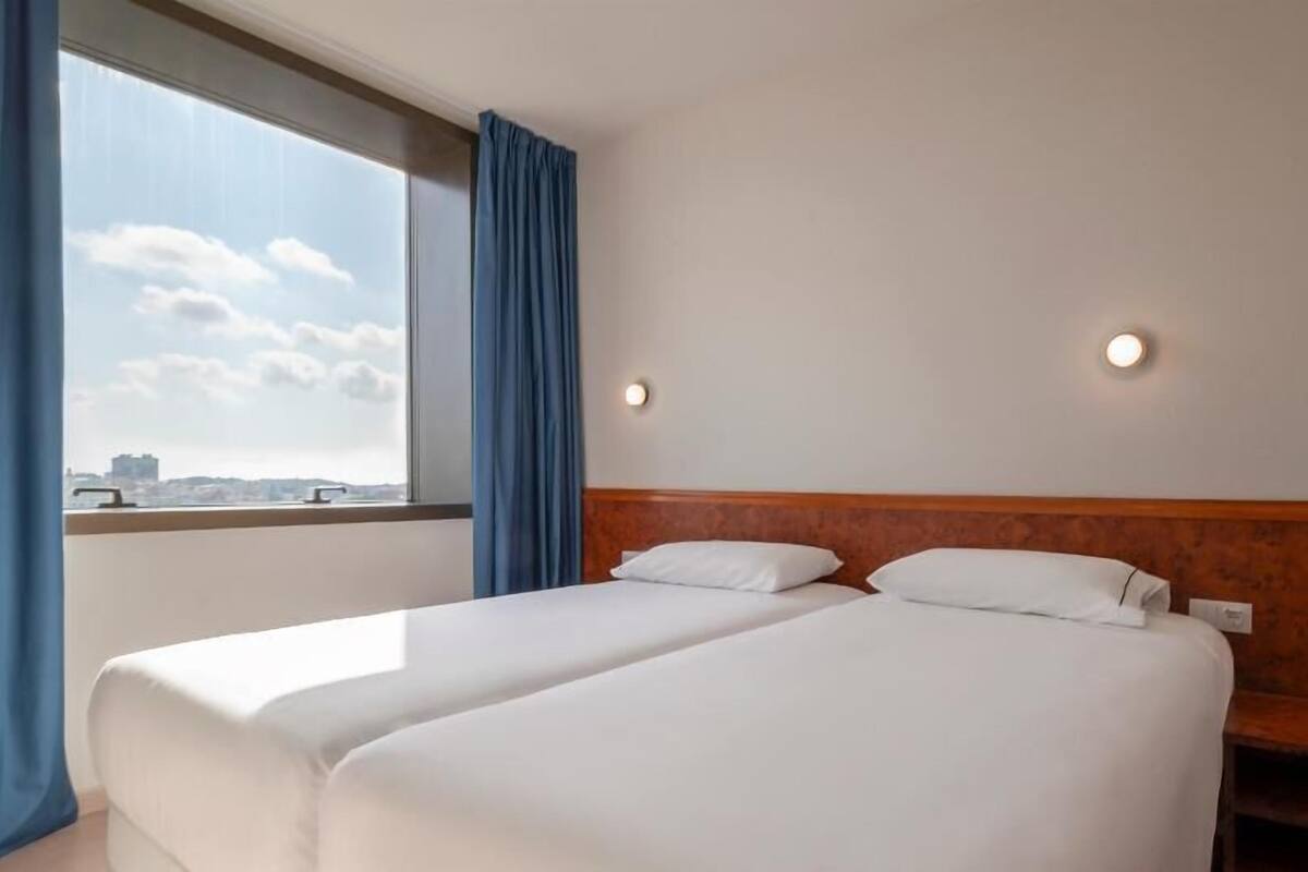 A hotel room with two beds, a large window with curtains, and a city view.
