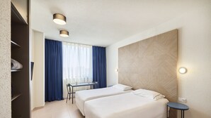 A hotel room with two beds, a desk, and a chair. There is a window with curtains, a wall-mounted light, and a wall with a geometric pattern. - Apartamentos Hesperia Sant Joan (Sant Joan Despi)